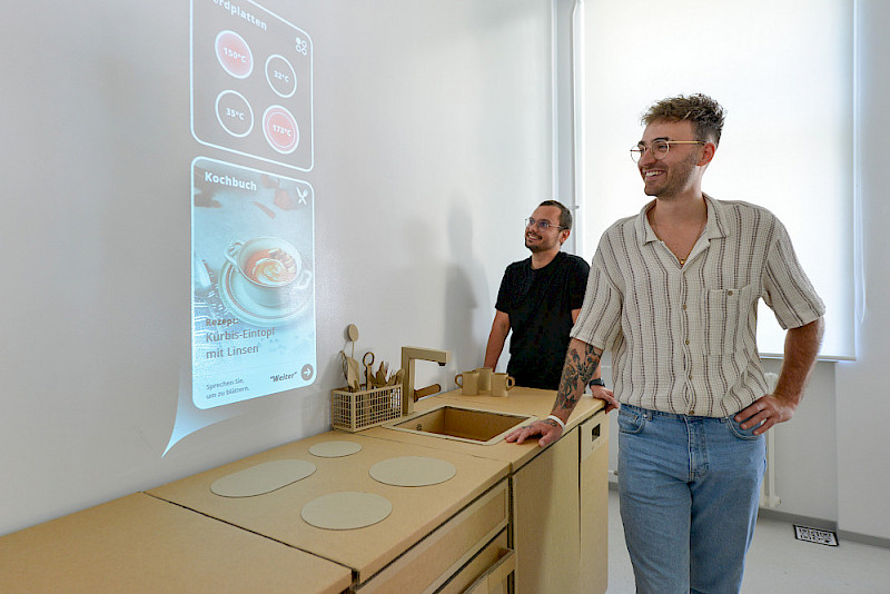Victor-Alexander Mahn (li.) und Martin Böhmer in der Modellküche - an der Wand ist neben Informationen zur Temperatur der Herdplatten ein Kochrezept eingeblendet.